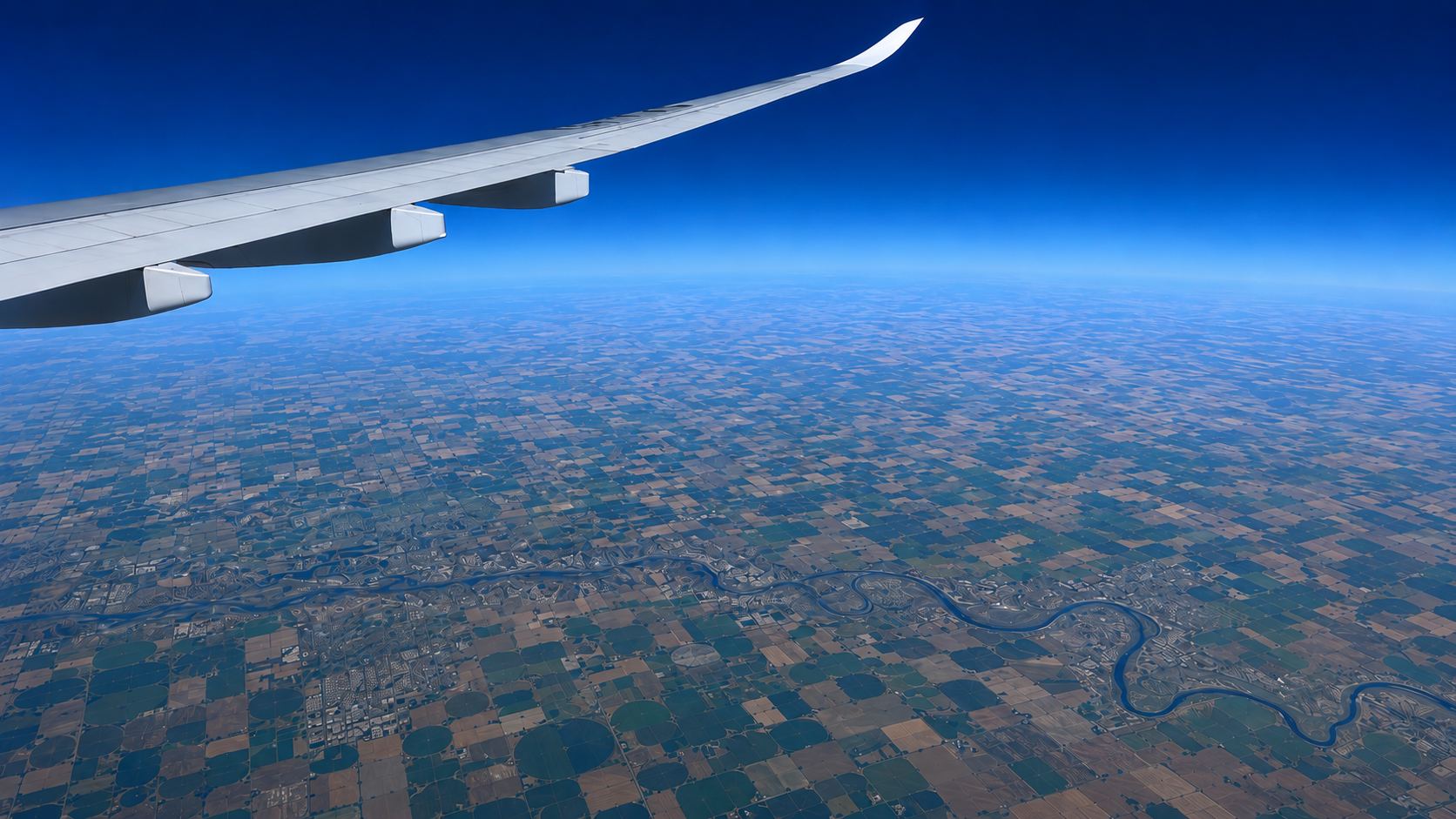 Campagna vista dal finestrino di un aereo: un mosaico di campi e un fiume sinuoso sotto un orizzonte blu intenso